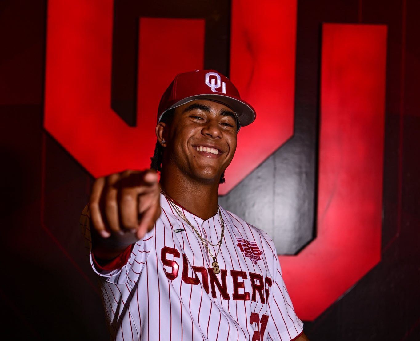 Malachi Witherspoon, standing in his Oklahoma Sooners baseball uniform in front of large OU letters, points toward the camera.